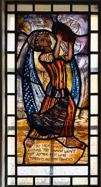 Stained glass window depicting a woman in traditional dress holding a tambourine, with a background of women and angels, and an inscription at the bottom that reads, 'In her hand and all the women went out after her with tambourines and with dances.'