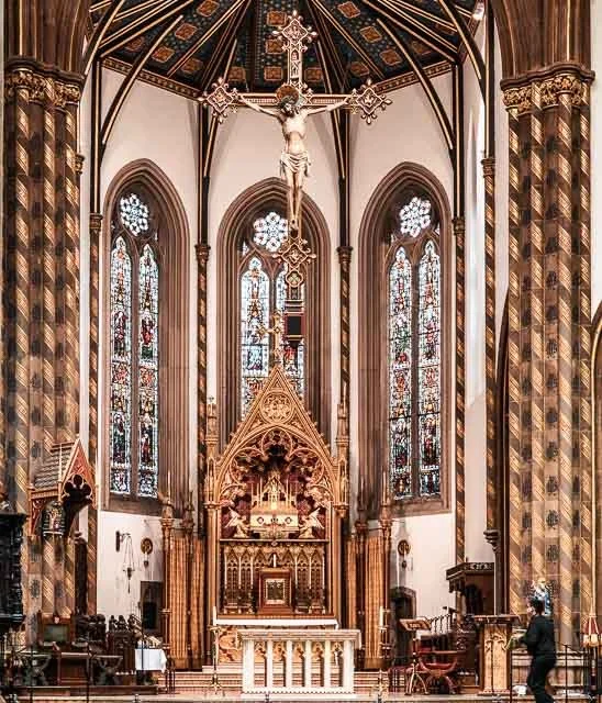 Interior of a Gothic-style church with a high vaulted ceiling, stained glass windows, a crucifix hanging above the altar, and elaborate woodwork.