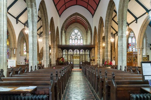 Interior of a historic church with wooden pews, stained glass windows, stone columns, and a high arched ceiling.