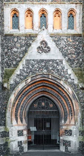 Close-up of a historic church entrance with a rounded archway framed by multiple layers of stone, topped by stained glass windows and a stone decorative emblem.