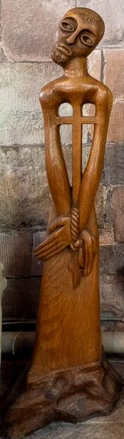 Wooden sculpture of a tall, slender figure with a skull-like head, elongated arms, and a cross-shaped opening in the chest, standing against a brick wall.