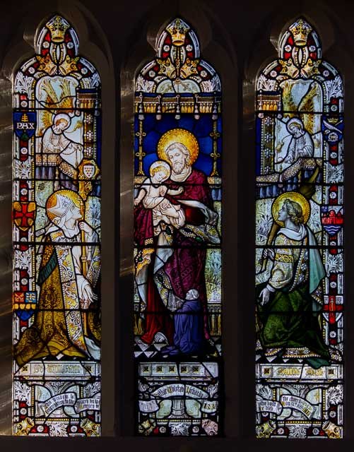Three stained glass windows in a church depicting religious figures. The middle window shows Jesus Christ holding a child, with a halo around his head. The left and right windows show saints or apostles with halos, holding scrolls or books.