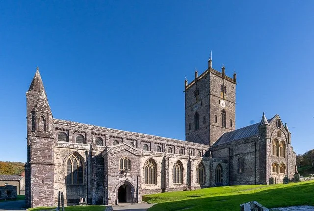 Ancient stone church with a tall bell tower, arched windows, and Gothic architecture, set against a clear blue sky with a green grassy area in the foreground.
