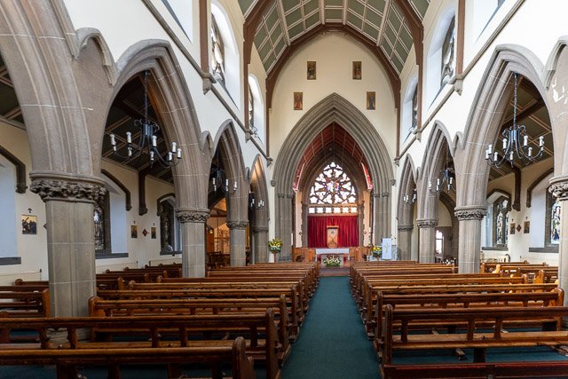Interior of a church with wooden pews, stone columns, and a stained glass window at the altar.