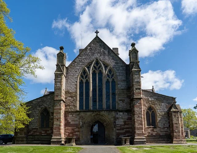 Front view of a historic stone church with arched windows, pointed gables, and decorative stonework, set against a partly cloudy sky, with green trees and lawn.