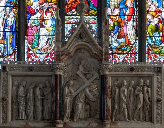 Stone carving depicting scenes from the Passion of Christ, with stained glass windows featuring biblical figures in the background.