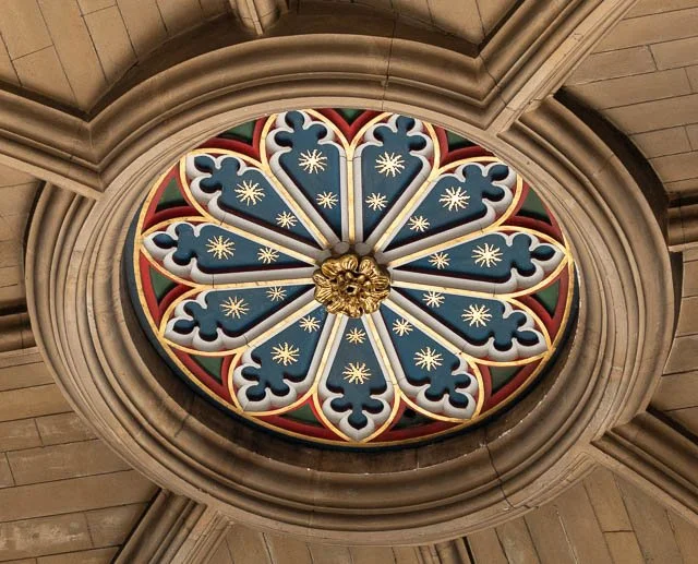 View of a decorative stained glass rose window with intricate patterns and a gold centerpiece, set in a stone ceiling.