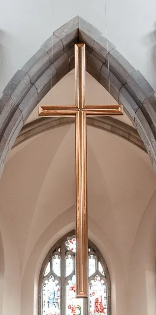 A large gold cross hanging from the ceiling inside a church with stone archways and stained glass windows.