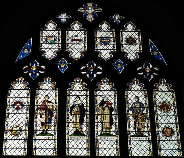 Colorful stained glass window depicting four historical figures dressed in period clothing, with intricate patterns and shields at the top.