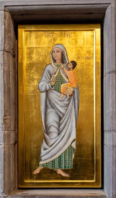 A religious icon painting of a woman holding a child, both dressed in robes, set in a golden background, framed within a stone structure.