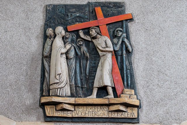 Wooden religious relief sculpture depicting Jesus carrying the cross, with several women and soldiers nearby, mounted on a wall with an inscription underneath that reads 'Jesus Speaks to the Daughters of Jerusalem.'