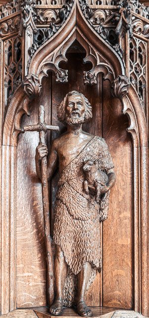 Wooden sculpture of a man with a beard holding a long staff and a small animal, standing in a decorative arched alcove.