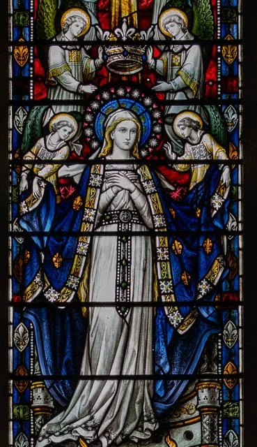 A stained glass window depicting a crowned woman in white and blue robes, surrounded by angels and religious symbols.