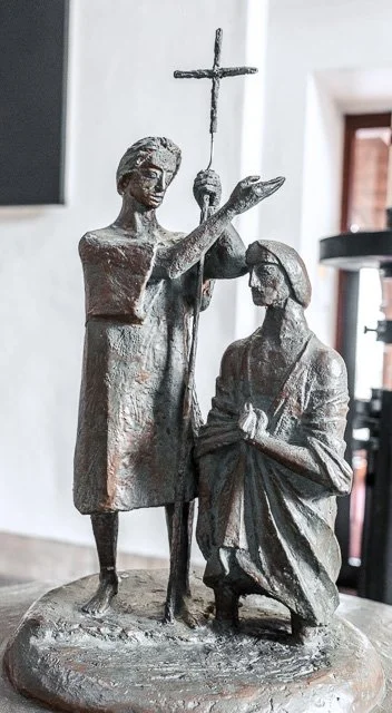 A metal sculpture depicting a woman holding a cross above a kneeling man. The woman stands confidently while the man kneels with hands clasped.