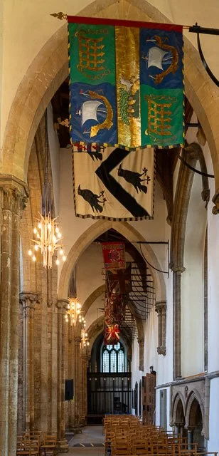 Interior view of a historic church with tall arched ceilings and stone columns. Banners hang from the ceiling, featuring coats of arms and heraldic symbols. Wooden pews line the aisle, leading towards an altar at the back.