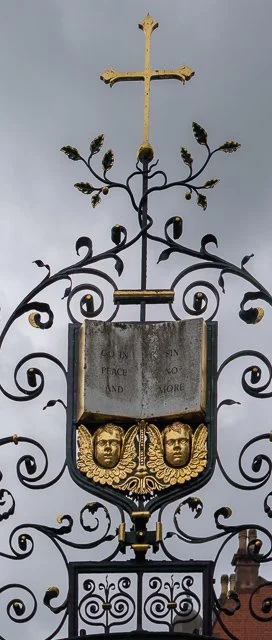A decorative metal sign with a cross on top, ornate gold details, and two face sculptures near the bottom, set against a cloudy sky.