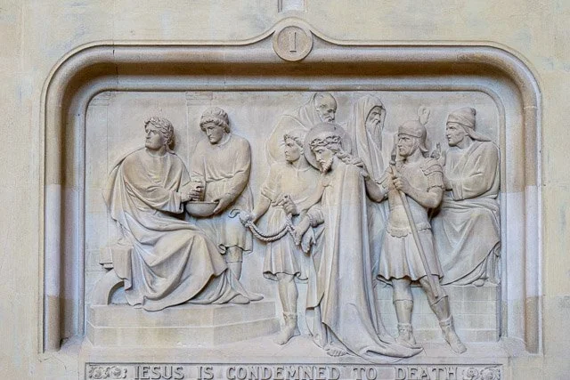 Relief sculpture depicting Jesus condemning a group of people, with some in robes and others in armor, on a stone wall.