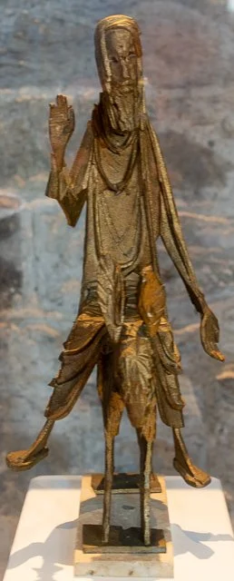 A carved wooden sculpture of a standing figure with a raised right hand, draped in clothing, mounted on a horse, displayed on a white platform.