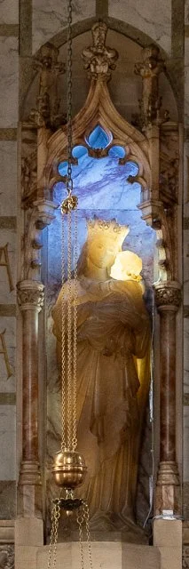 A religious statue of a saint, adorned with a crown and robes, encased in a decorative wooden and glass shrine with ornate carvings and a chain hanging in the foreground.