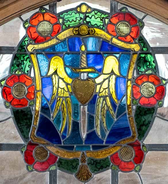 A stained glass window featuring a shield with a sword, a heart, and a pair of yellow wings, surrounded by a floral border of red poppies and green leaves.