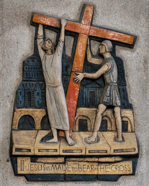 Wooden relief sculpture depicting Jesus carrying the cross with a Roman soldier beside him, set against a cityscape background, with an inscription that reads 'Jesus is made to bear the cross' at the bottom.