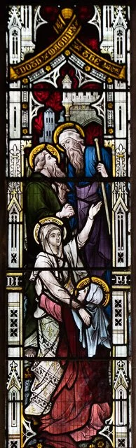 A stained glass window depicting a religious scene with four figures, including an elderly man with a beard and a woman with a halo, all dressed in traditional robes, surrounded by Gothic architectural elements and inscriptions.