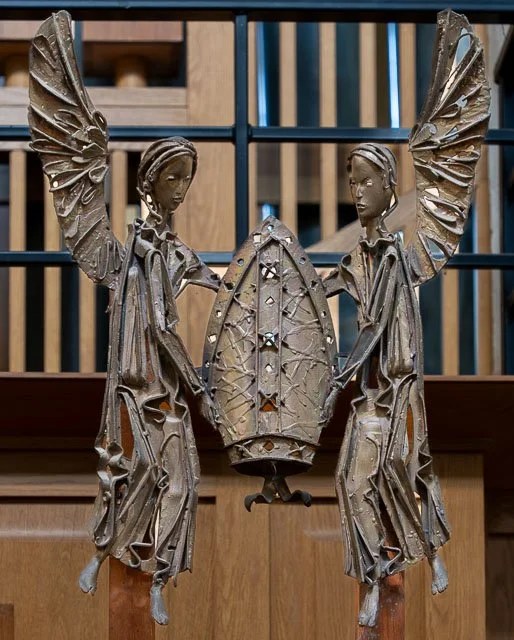 A metallic sculpture of two angels holding a shield, with wings extended, placed in front of a wooden background.