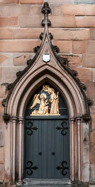 A gothic-style arched church door with decorative stone framing and ornate detailing, set in a brick wall.