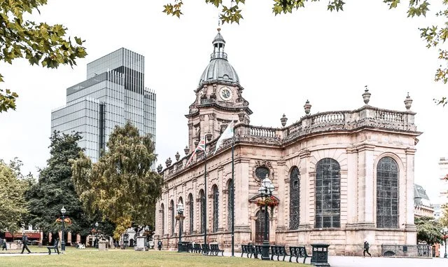 Historic stone building with a clock tower and domed top, surrounded by trees, park benches, and modern glass skyscrapers in the background.