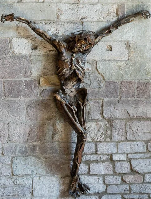 A sculpture of a crucifix made from twisted and arranged wooden pieces, mounted on a brick wall.