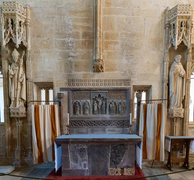 Interior of a historic church or cathedral with an altar, surrounded by ornate stone statues on tall pedestals, framed by decorative curtains on either side.