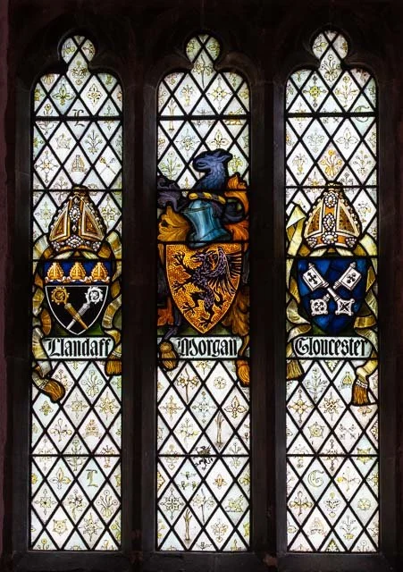 Three stained glass windows with coat of arms and heraldic symbols, featuring a dragon in the center holding a shield, flanked by two other shields with crown and crossed swords, and banners with names Southampton, Arundel, and Gloucester.