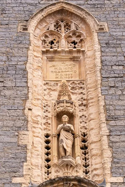 Stone Gothic-style wall with a niche housing a statue of a person holding an object, and an inscription above.
