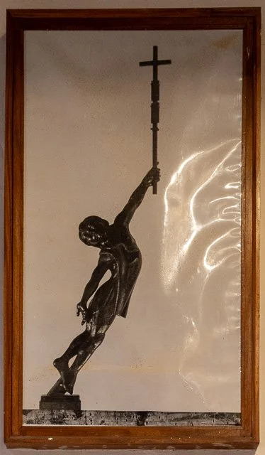 Religious sculpture of Jesus Christ on the cross with a long vertical cross above him, mounted inside a wooden frame.