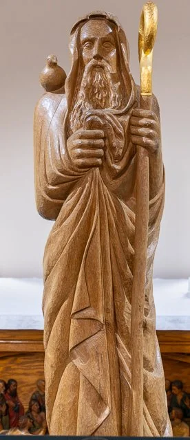 Wooden sculpture of a bearded man holding a staff, with a backpack on his back and a small bird perched on his shoulder.