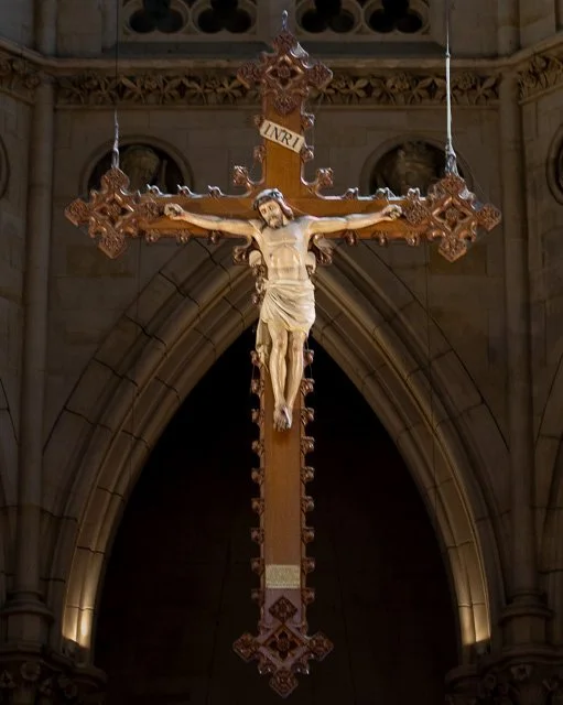 A crucifix with Jesus Christ on the cross hanging inside a church, with arches and intricate architectural details in the background.