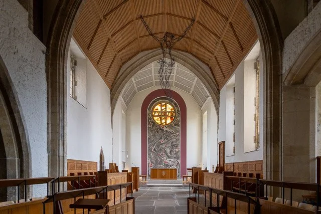 Interior of a church with a wooden ceiling, stone walls, wooden pews, a marble altar at the front, and a large circular stained glass window at the back.