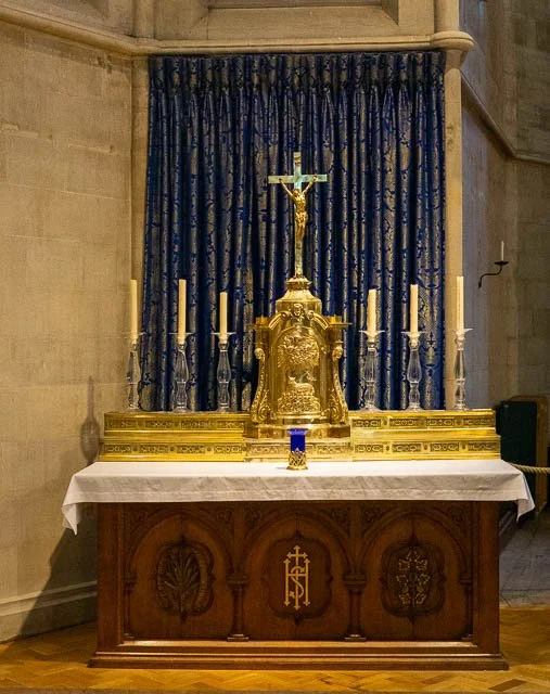 Gold crucifix with Jesus on the cross, surrounded by seven tall candlesticks with candles, placed on an ornate gold altar with a white cloth, in front of a blue curtain with a gold pattern.
