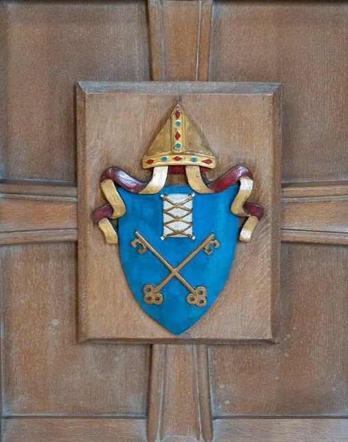 Colorful coat of arms featuring a blue shield with two crossed gold keys, a gold crown at the top, and a gold and red helmet on a rectangular wooden plaque.