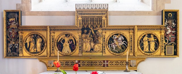 An ornate gold altarpiece with religious paintings and carvings, flanked by candles and flowers.