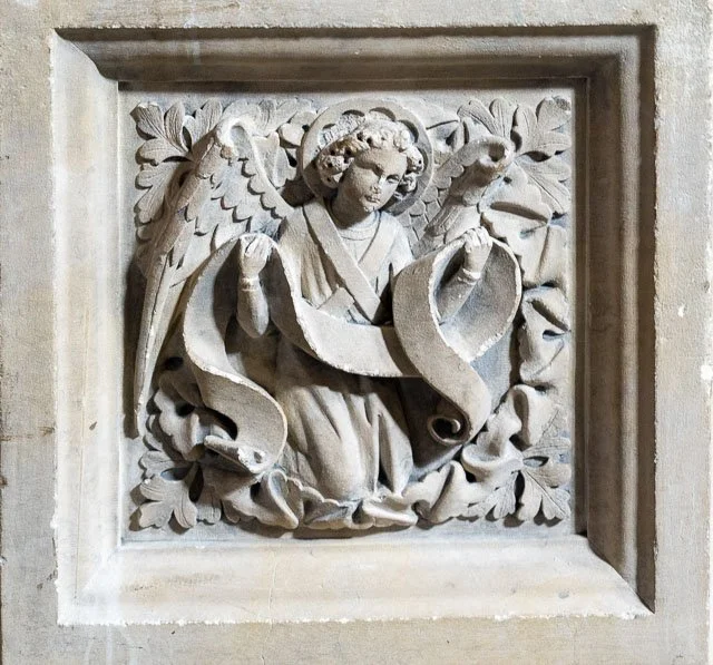 Carved stone relief of an angel with wings, holding a shield, surrounded by leaves.