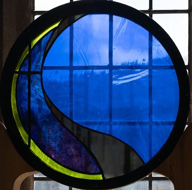 Colorful stained glass window featuring blue, green, purple, and black glass pieces in an abstract circular design.