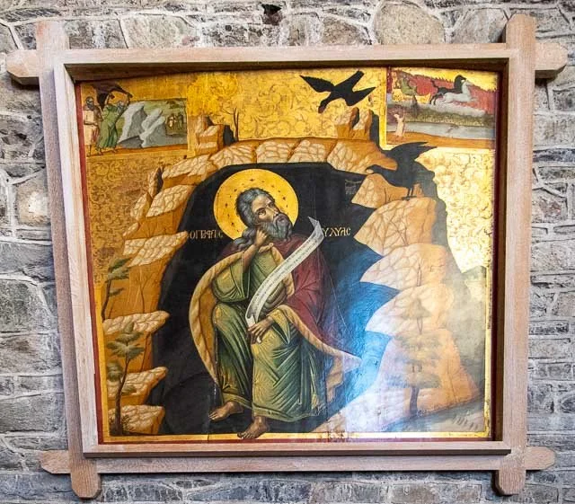 A religious icon depicting a saint with a halo, holding a scroll and a pen, surrounded by rocky terrain, trees, and flying birds.
