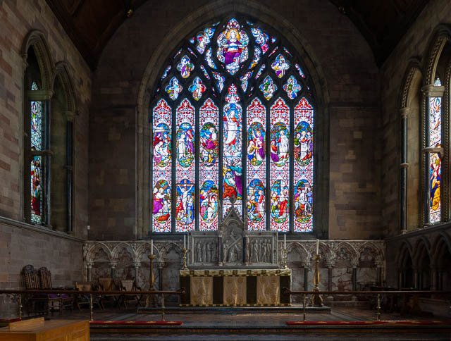 Interior of a church with a large stained glass window behind the altar, depicting religious scenes and figures, flanked by smaller stained glass windows on either side.