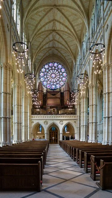 Interior of a cathedral with vaulted ceiling, rows of wooden pews, wall-mounted light fixtures, and a large stained glass rose window at the front.