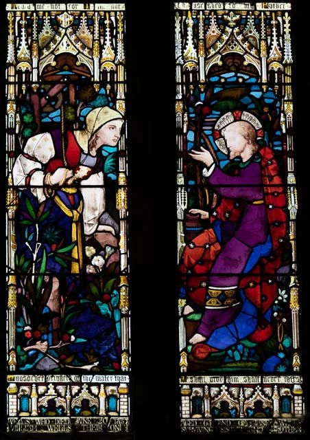 Colorful stained glass windows depicting a woman guiding a man who appears to be injured or in need, with decorative Gothic architectural elements in the background.