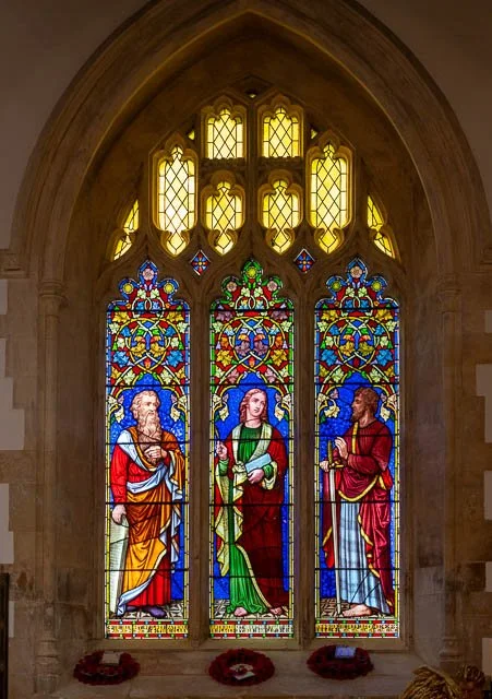 Colorful stained glass window with three biblical figures inside a church, with decorative arches and flower arrangements at the base.