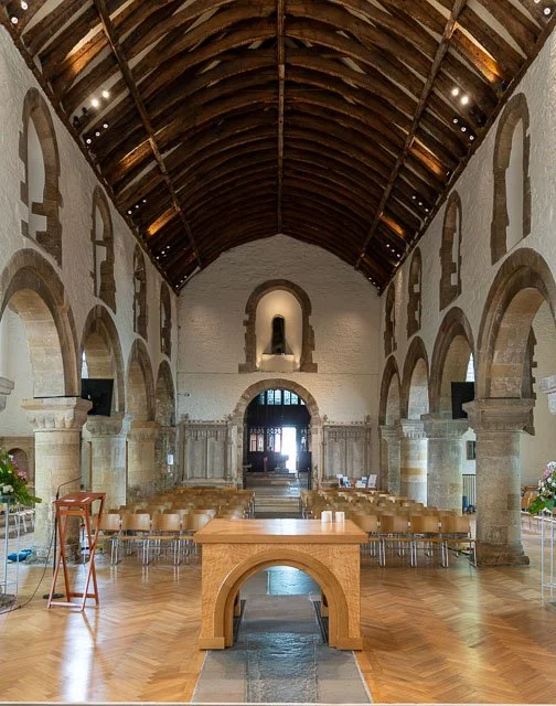 Interior of a historic church or chapel with a high wooden vaulted ceiling, stone arches, and rows of wooden chairs facing the altar.