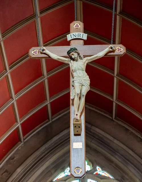 Wooden crucifix with Jesus Christ figure hanging inside a church, with a stained-glass window at the bottom