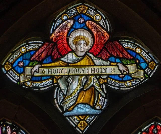 Stained glass window depicting an angel with red wings holding a banner that says 'HOLY HOLY HOLY' in a church setting.